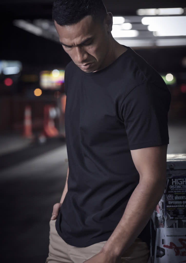 A black men's t-shirt is displayed on a model, set against a blurred urban background with street signs.