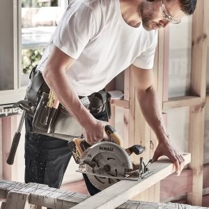 A man in a white T-shirt uses a circular saw on a wooden plank, wearing safety glasses and noise-cancelling headphones.