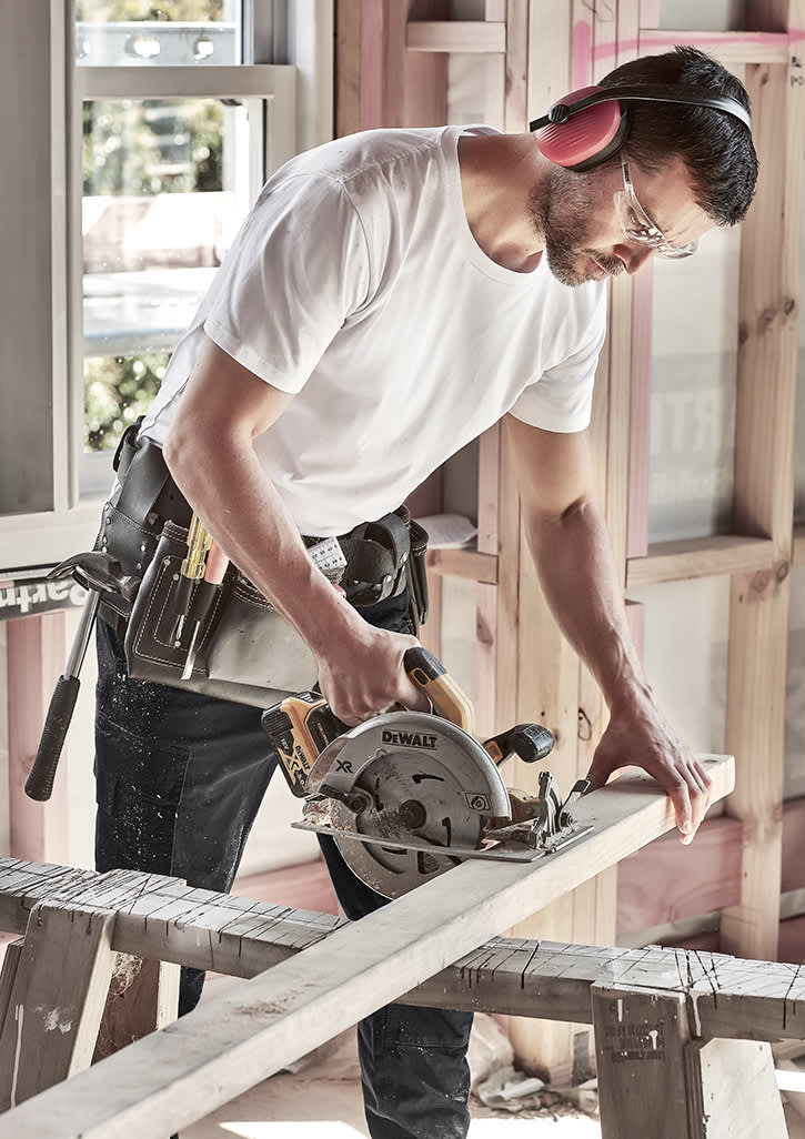 A man in a white T-shirt uses a circular saw on a wooden plank, wearing safety glasses and noise-cancelling headphones.