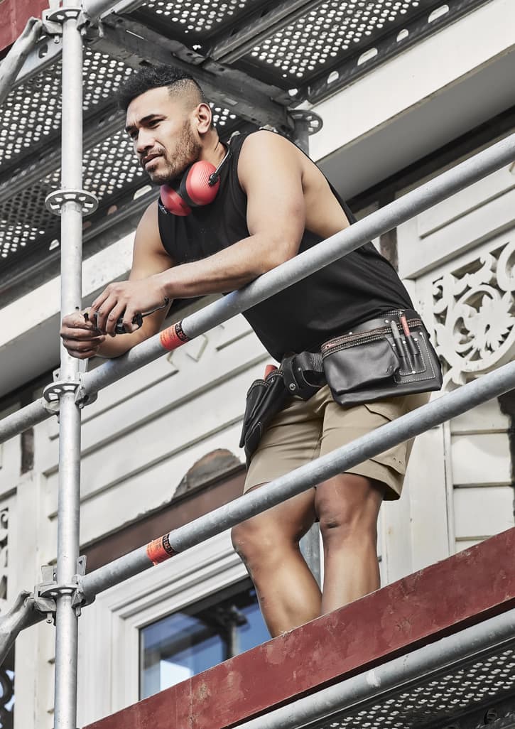 A man wearing a black sleeveless tee, shorts, and a tool belt stands on scaffolding, with headphones around his neck.