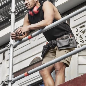 A man in a black sleeveless tee and shorts stands on scaffolding, wearing headphones and a tool belt.