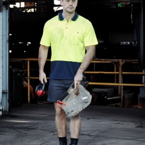 A man wearing a high-vis yellow and navy polo shirt, shorts, and a hard hat, holding work gear in a warehouse setting.