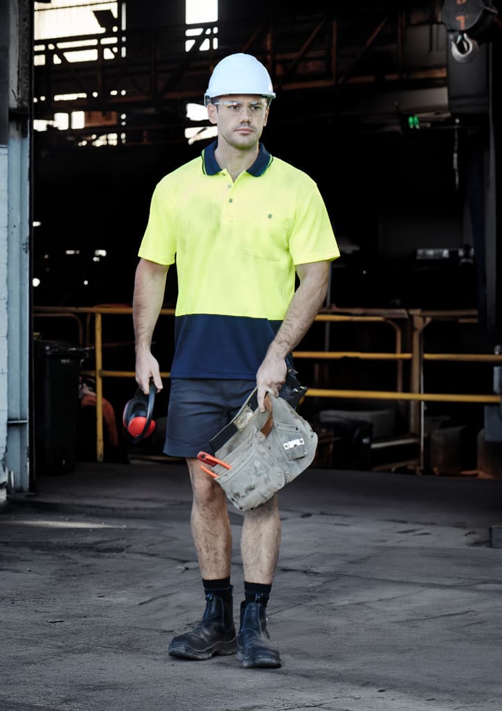 A man wearing a high-vis yellow and navy polo shirt, shorts, and a hard hat, holding work gear in a warehouse setting.