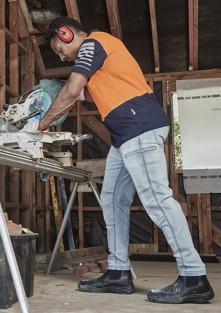 A man wearing an orange and navy short sleeve polo shirt works with a saw in a workshop setting.