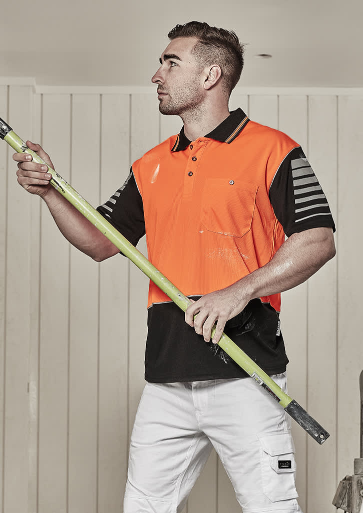 A man wearing an orange and black short sleeve polo shirt stands holding a green pole. The shirt has a logo.