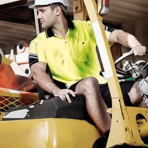 A man wearing a yellow and navy hi-vis short sleeve polo shirt operates a forklift, wearing a safety helmet.