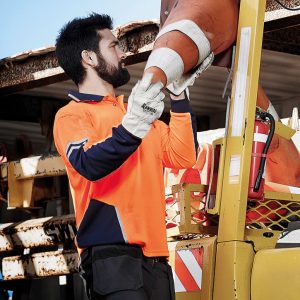 A man in an orange and navy long sleeve polo lifts a traffic cone near a yellow vehicle. The scene is outdoors.