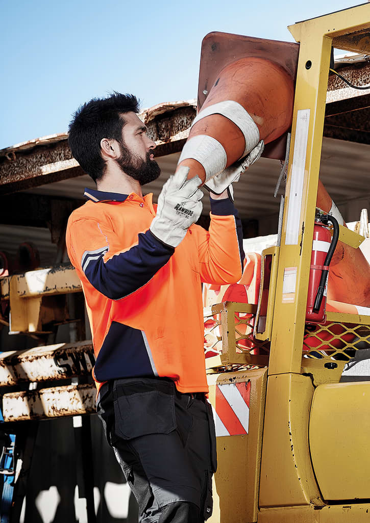 A man in an orange and navy long sleeve polo lifts a traffic cone near a yellow vehicle. The scene is outdoors.