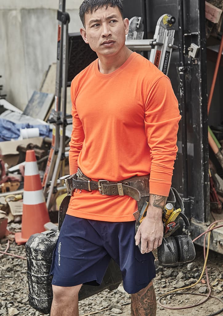 An orange long sleeve safety tee is worn by a man with a tool belt, set against a cluttered worksite background.