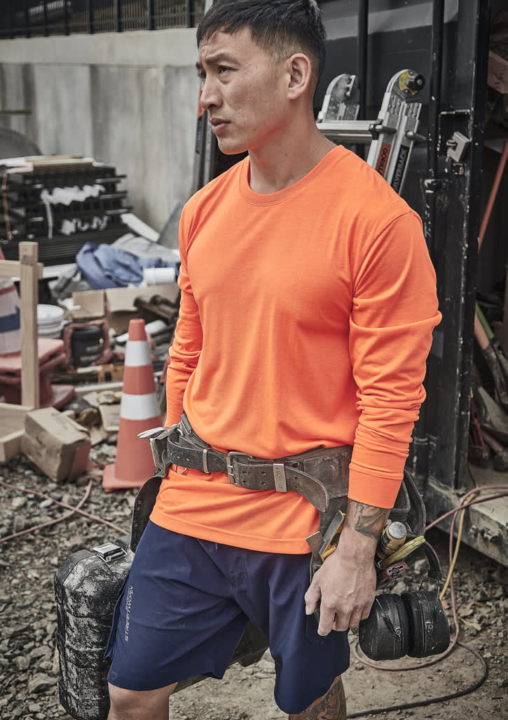 A man wearing an orange long sleeve high visibility tee is standing at a construction site, equipped with a tool belt.