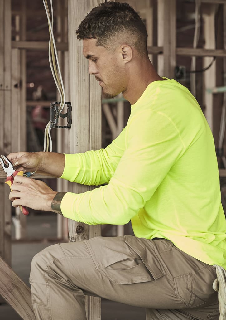 A man works in a construction setting, wearing a neon yellow long sleeve tee and beige cargo pants, using pliers.