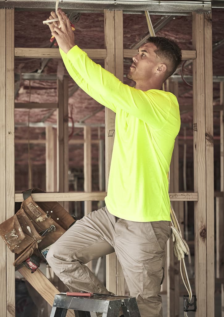 A man in a high visibility long sleeve yellow tee works on a ladder in a construction setting. The shirt has a logo.