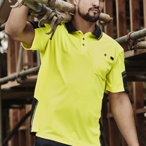 A man in a bright yellow short sleeve polo with black accents, carrying scaffolding pipes at a construction site.