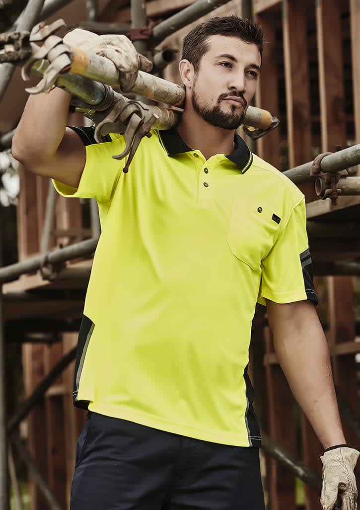 A man in a bright yellow short sleeve polo with black accents, carrying scaffolding pipes at a construction site.
