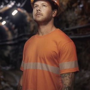 A man wearing an orange hi-vis segmented tape cotton tee shirt and a safety helmet with headlamp, standing in a dimly lit area.
