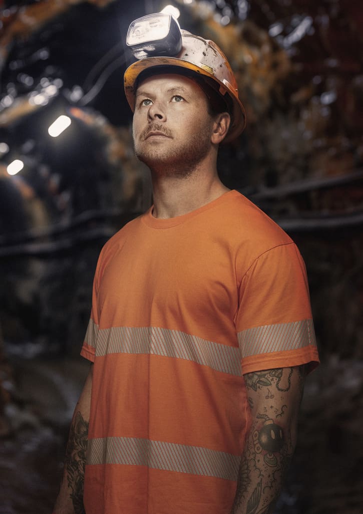 A man wearing an orange hi-vis segmented tape cotton tee shirt and a safety helmet with headlamp, standing in a dimly lit area.