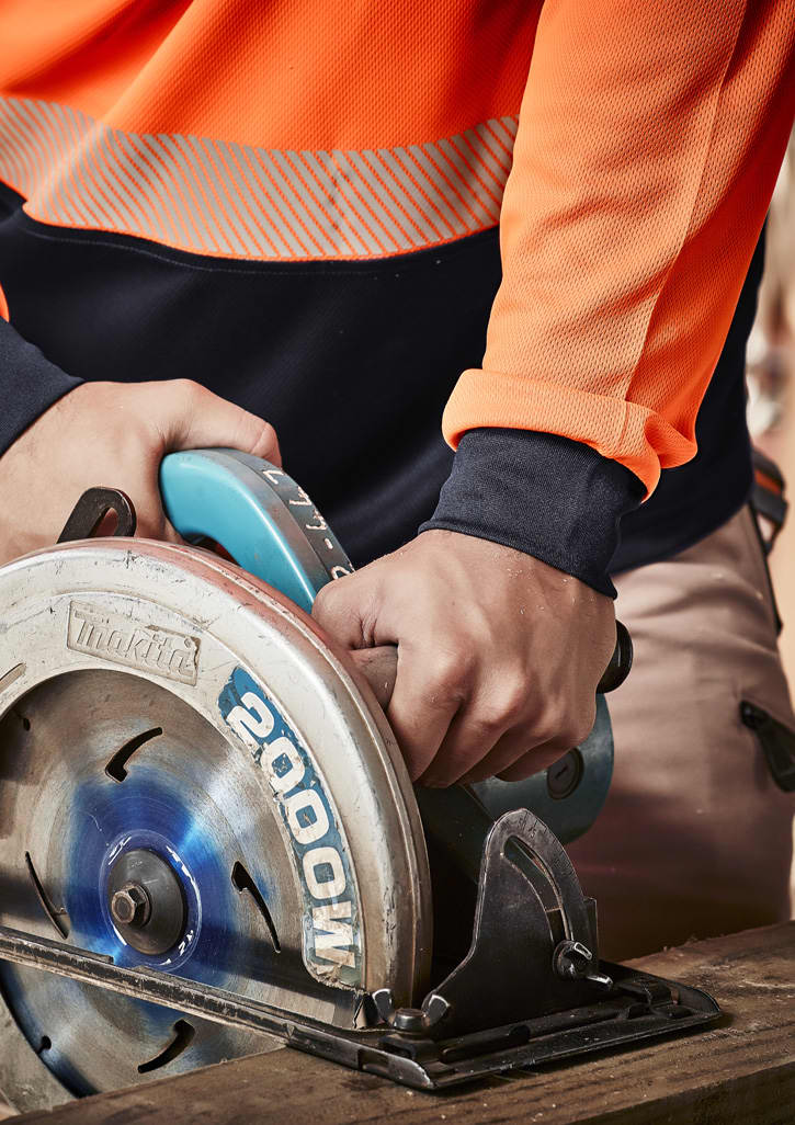 A close-up of hands gripping a circular saw, wearing an orange and navy long sleeve work polo shirt.
