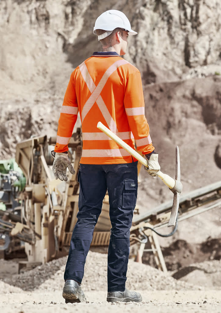 A worker in an orange long sleeve polo shirt with reflective stripes, holding a pickaxe, facing away from the camera.