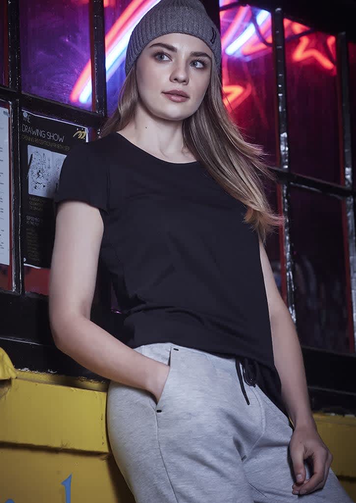 A woman in a black t-shirt and grey pants leans against a wall, wearing a beanie. The background features neon lights.