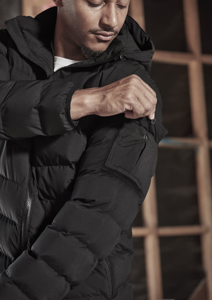 A black unisex puffer jacket with a hood, featuring a zip-up front and a pocket on the arm.
