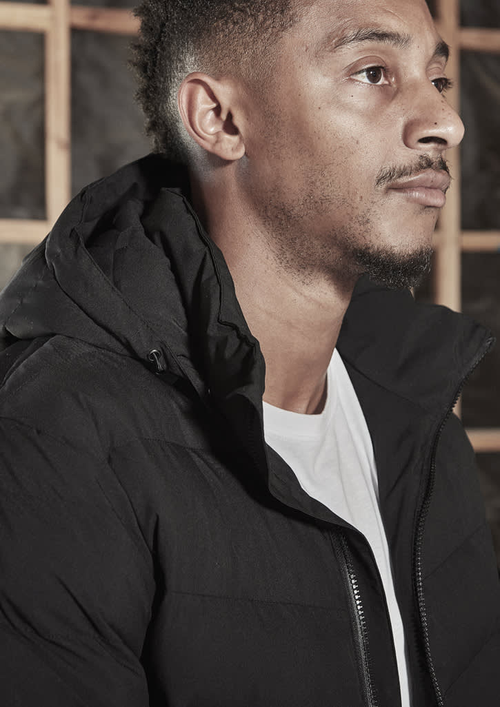 A black hooded puffer jacket, featuring a zipper and a discreet logo, worn by a model.