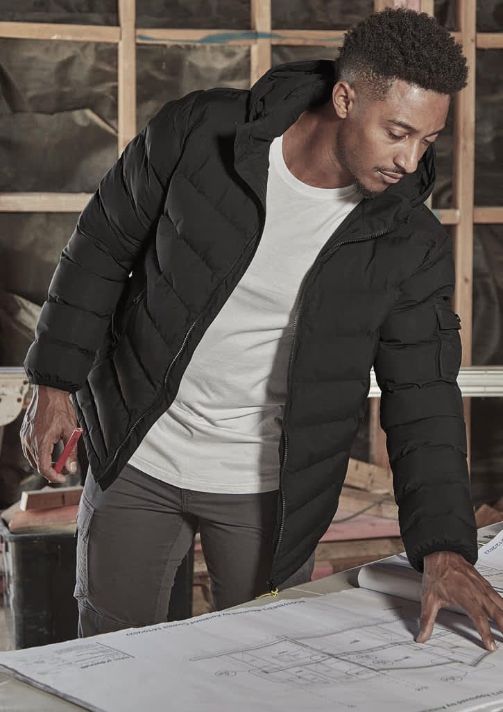 A black unisex puffer jacket with a hood, worn by a person examining blueprints indoors. The jacket has a logo.
