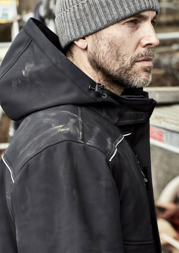 A black unisex softshell jacket with a hood, worn by a man with a grey beanie, features a logo and some dirt stains.