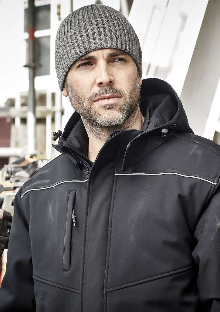A man in a black softshell jacket and grey beanie, standing in an outdoor setting with construction in the background.