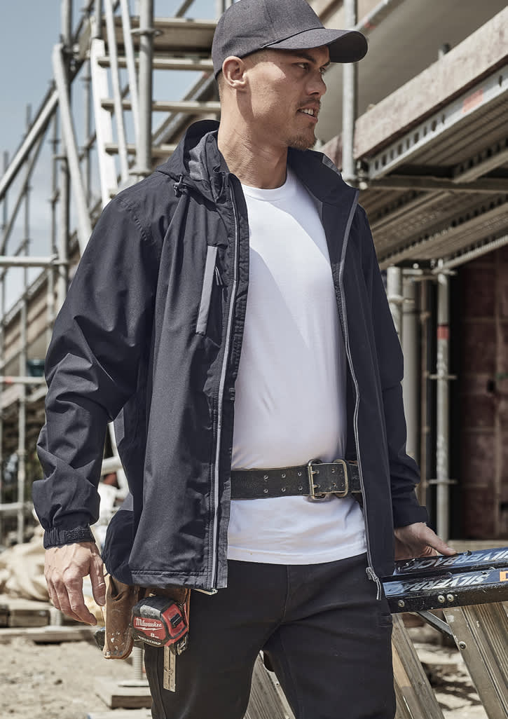 A man in a black packable rain jacket stands at a construction site, wearing a cap and tool belt.