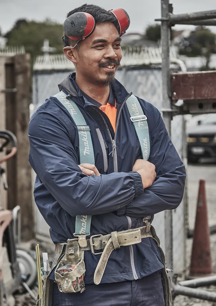 A man in a navy blue packable rain jacket stands with arms crossed, wearing earmuffs and a safety harness.