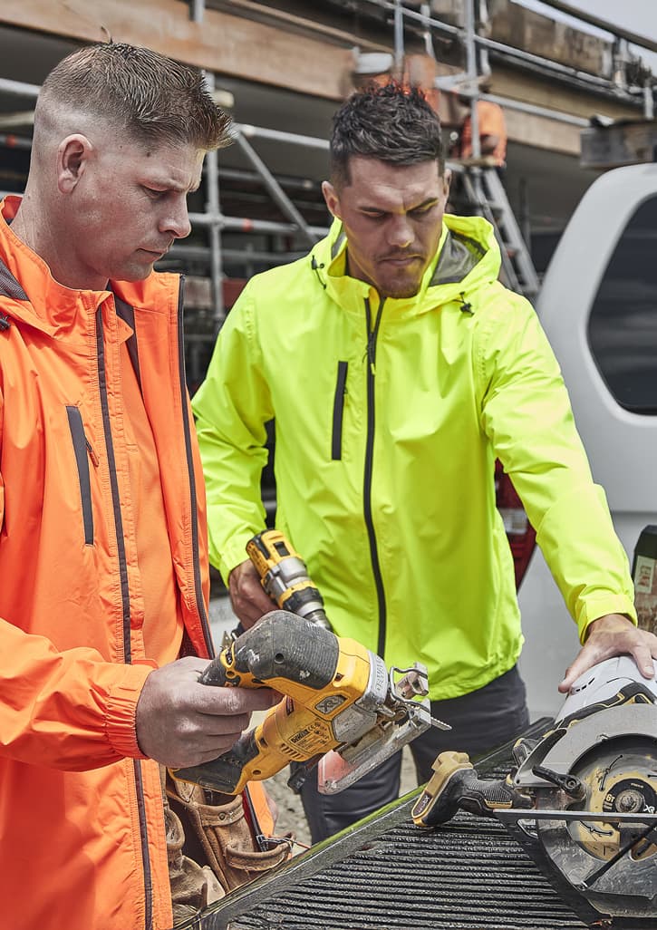 Two men in bright orange and neon yellow rain jackets work on a construction site, using power tools. Both jackets have logos.