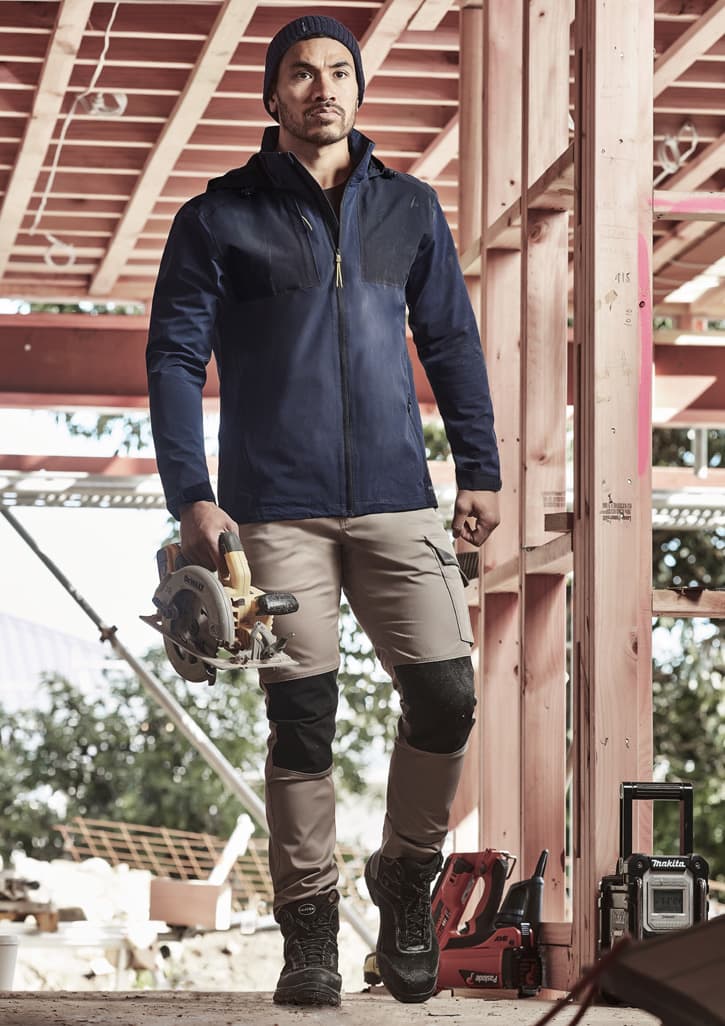 A man wearing a navy waterproof jacket stands on a construction site, holding a saw. He pairs it with beige pants and boots.