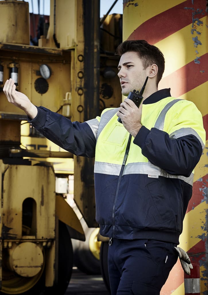 A man in a hi-vis quilted flying jacket, featuring yellow and navy colours, standing with a radio in a work environment.