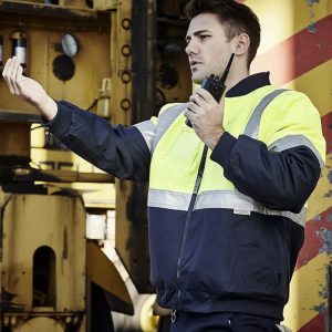 A man in a hi-vis yellow and navy quilted jacket uses a walkie-talkie against an industrial background.