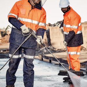 Two men in orange high-visibility jackets and helmets, working with tools on a construction site.