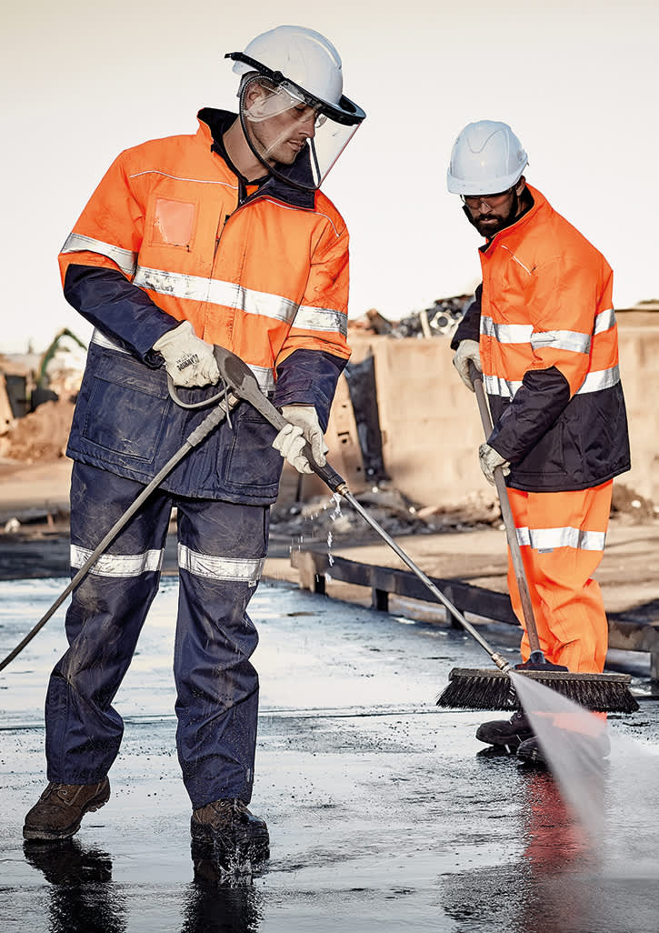 Two men in orange high-visibility jackets and helmets, working with tools on a construction site.