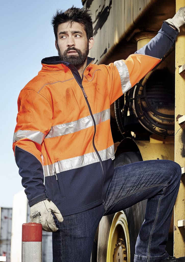 A unisex hi-vis softshell jacket in orange and navy, featuring reflective strips, worn by a person leaning against machinery.