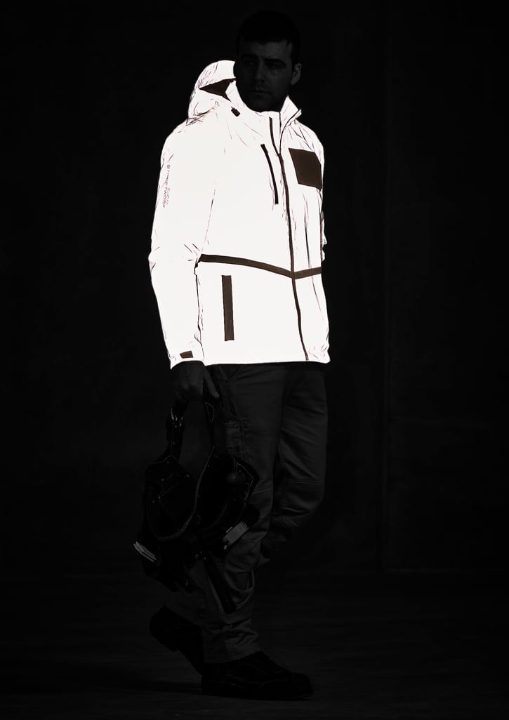 A reflective waterproof jacket in white, featuring a hood, zip pockets, and a logo, displayed on a model in a dark setting.
