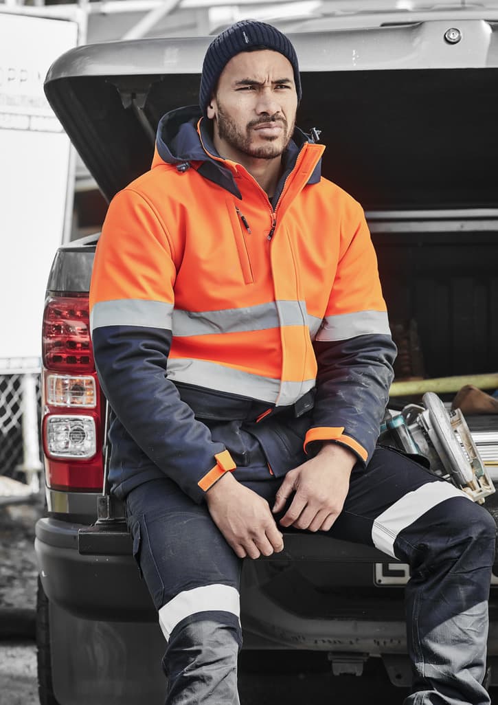 A man wearing an orange and navy unisex hi-vis softshell jacket sits on the back of a vehicle.