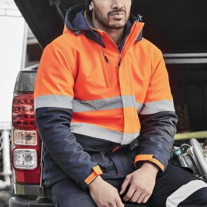 A unisex hi-vis softshell jacket in orange and navy, featuring reflective stripes, worn by a person sitting on a vehicle.