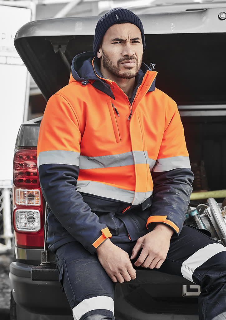 A unisex hi-vis softshell jacket in orange and navy, featuring reflective stripes, worn by a person sitting on a vehicle.