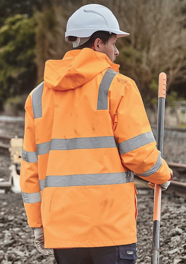 A person wearing an orange waterproof jacket with reflective stripes and a white hard hat, standing near railway tracks.