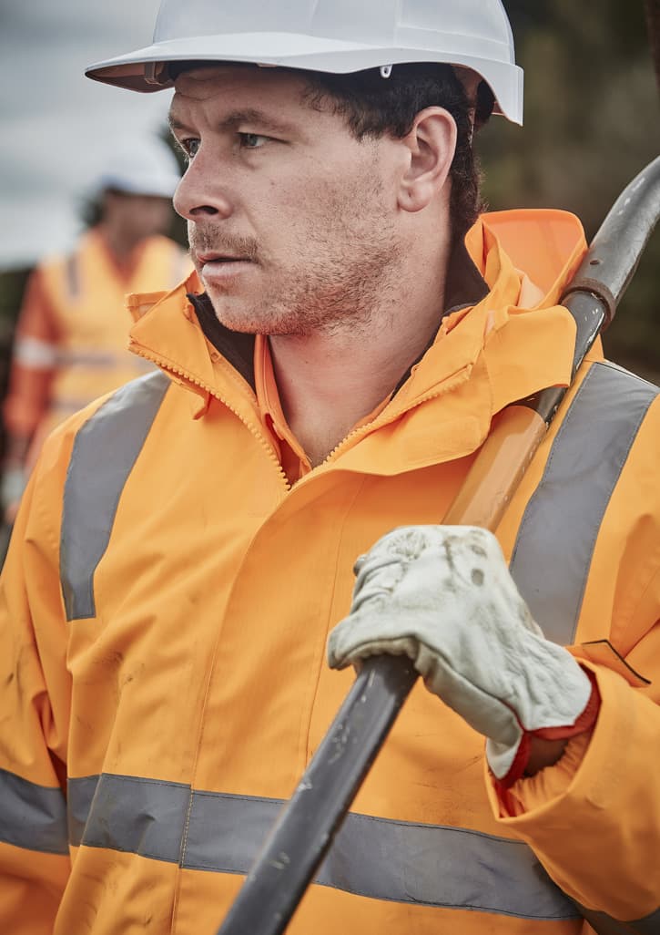 A man wearing an orange waterproof jacket with reflective strips, gloves, and a safety helmet, holding a tool.