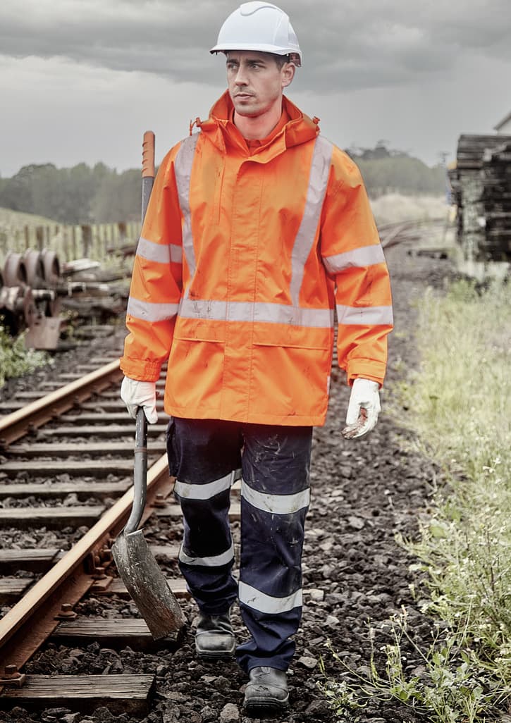 A man wearing an orange waterproof jacket and navy trousers walks along railway tracks, holding a tool. The jacket features reflective stripes.