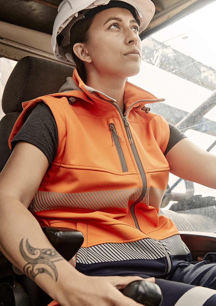 An orange softshell jacket with reflective detailing, worn by a woman, seated in a vehicle with a hard hat.
