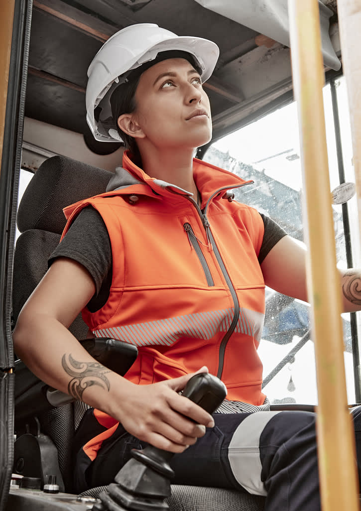 A woman wearing an orange softshell jacket with reflective strips, seated in a construction vehicle, wearing a hard hat.