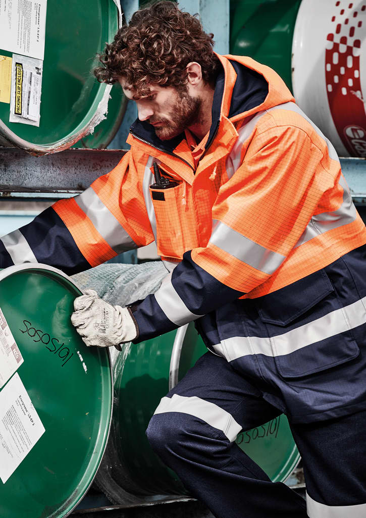 A man in an orange flame-resistant jacket adjusts a green barrel, wearing gloves and dark trousers.