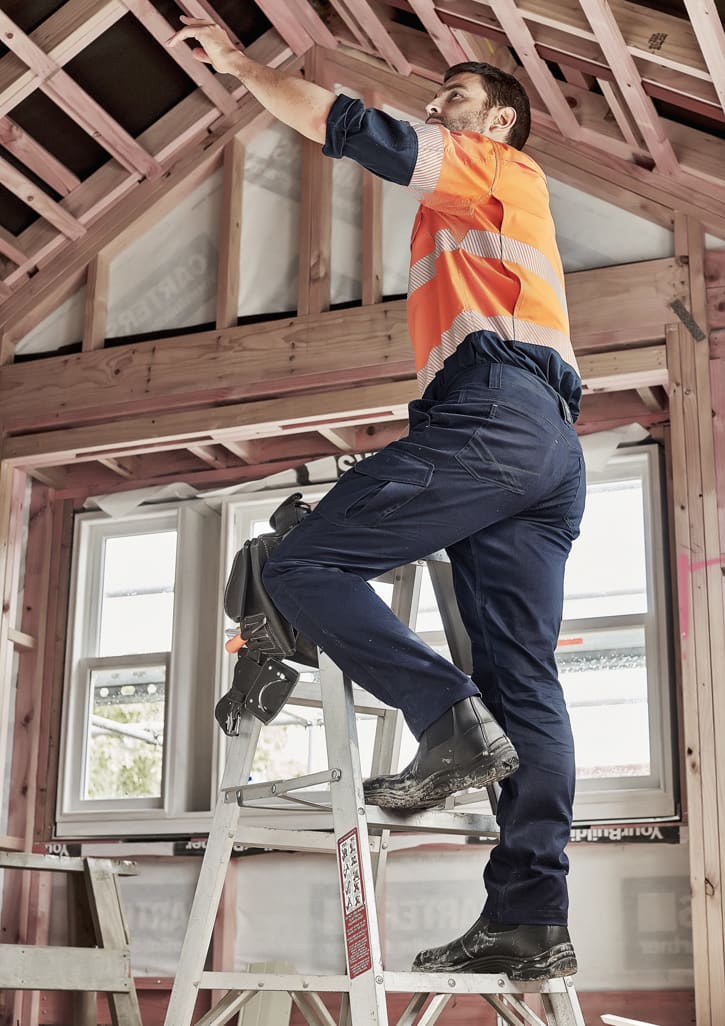A man in orange and grey high-visibility workwear wears navy cargo pants while standing on a ladder.