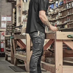 A man in black cargo pants and a black shirt standing at a workbench in a workshop, focused on a task.