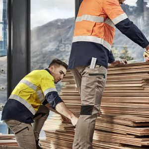 Two men wearing high-visibility work shirts in orange and yellow, with stretch pants, are arranging wooden boards indoors.